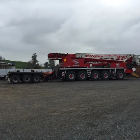 Red multi-axle mobile crane coupled to a low trailer on a gravel yard, with workers at the join between the two under an overcast sky
