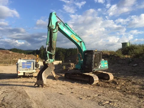 Green tracked excavator with a bucket and thumb attachment on an earthworks site, with a field service truck parked beside it