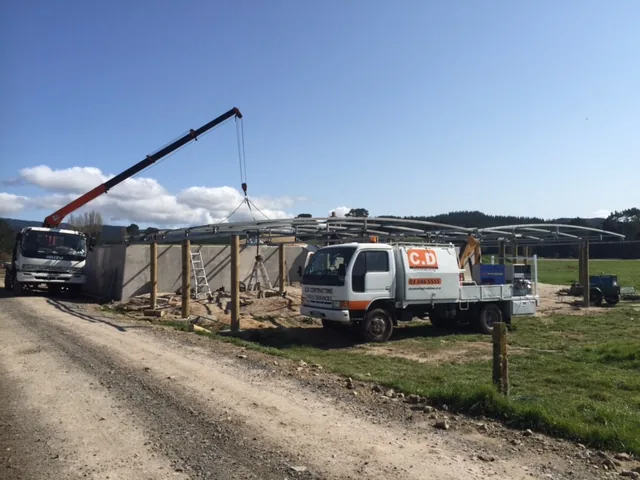 Truck-mounted crane lifting a curved steel roof frame onto timber poles on a rural site, with a white C.D service truck parked in front