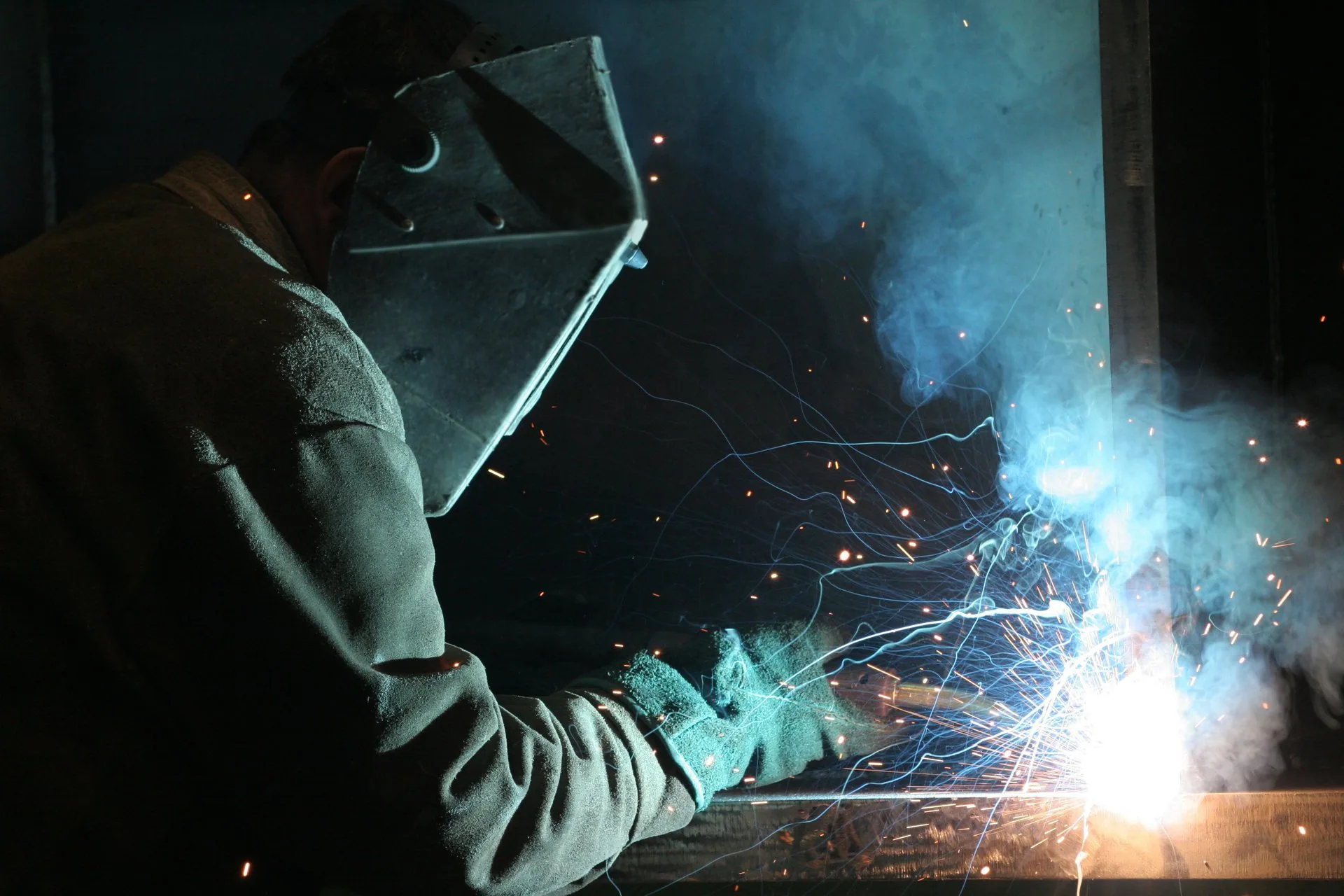 Stock photograph of a welder in a helmet and gloves arc welding in a dark workshop, lit blue by the arc with sparks flying